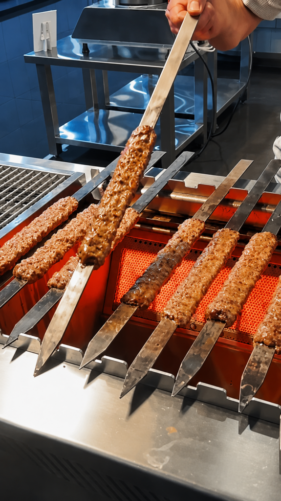Skewers of kebab meat grilling on a commercial grill as a cook lifts one with a metal spatula.